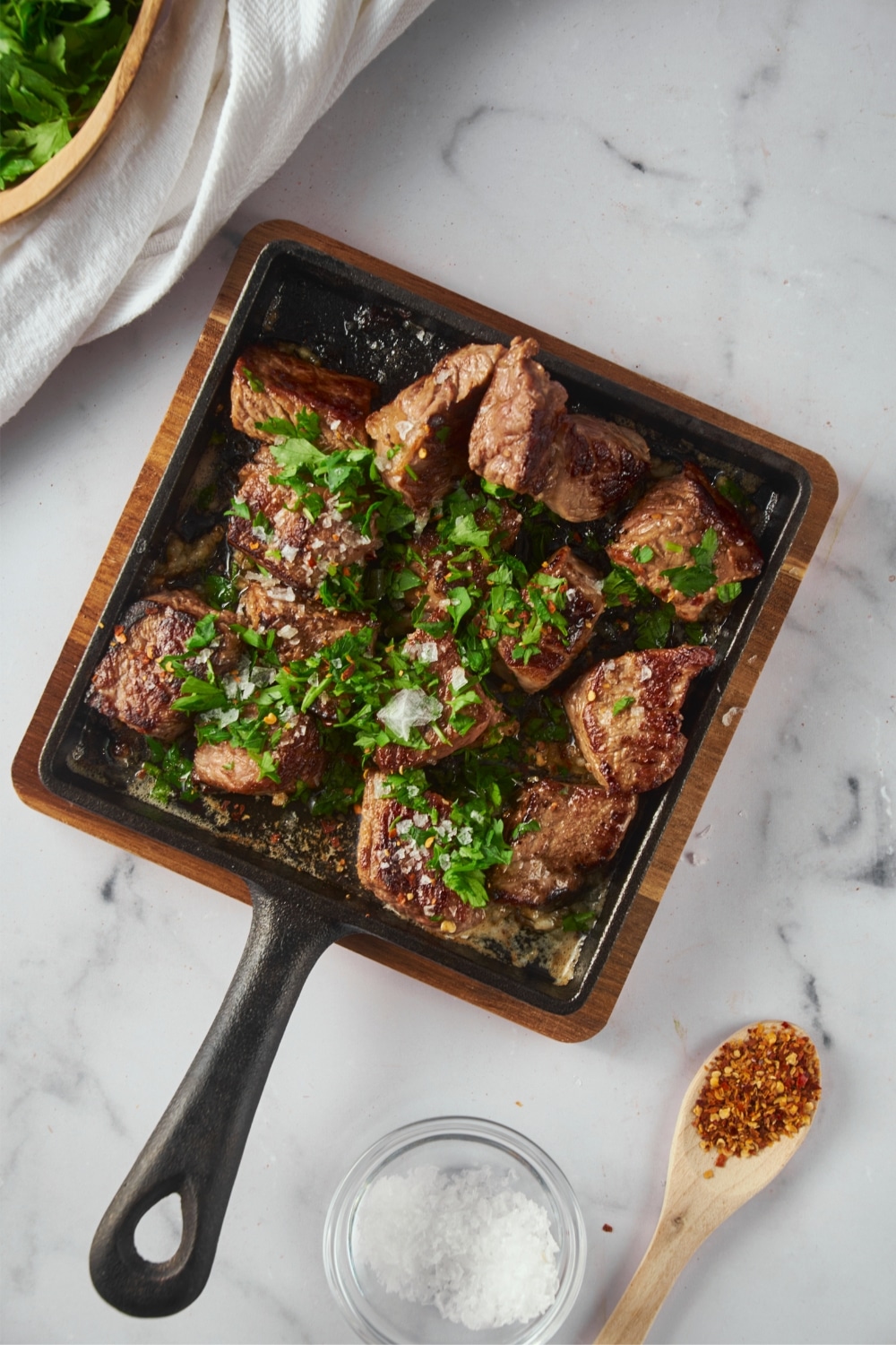 Top view of garlic steak bites garnished with parsley on a square cast iron pan next to a small bowl of salt and a spoonful of red pepper flakes.