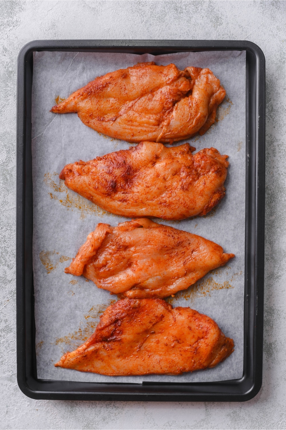 Seasoned and flattened thin chicken breasts on a parchment paper lined baking sheet.