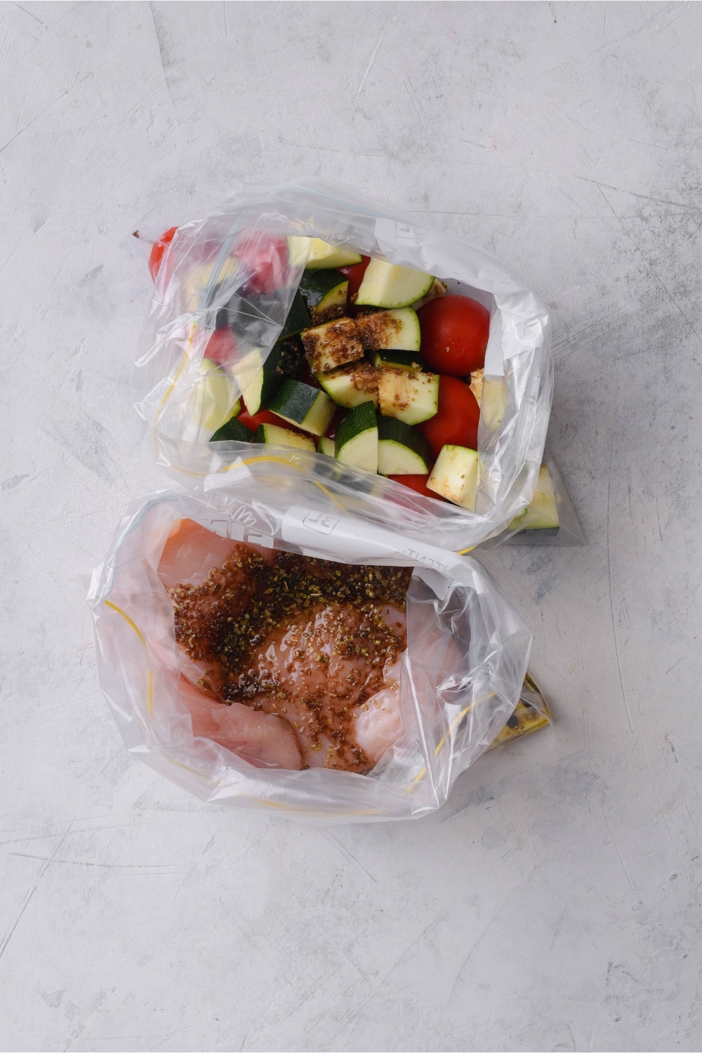 Two plastic bags, one filled with seasoned tomatoes and zucchini, the other filled with seasoned chicken.