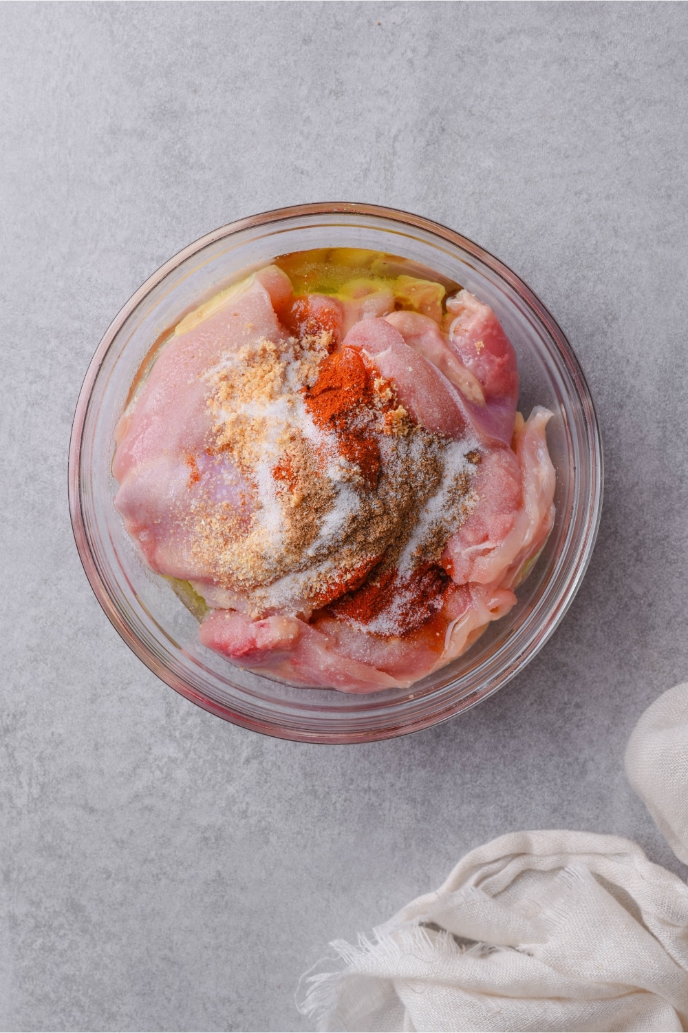 A glass bowl of raw boneless skinless chicken thighs with seasoning and oil.