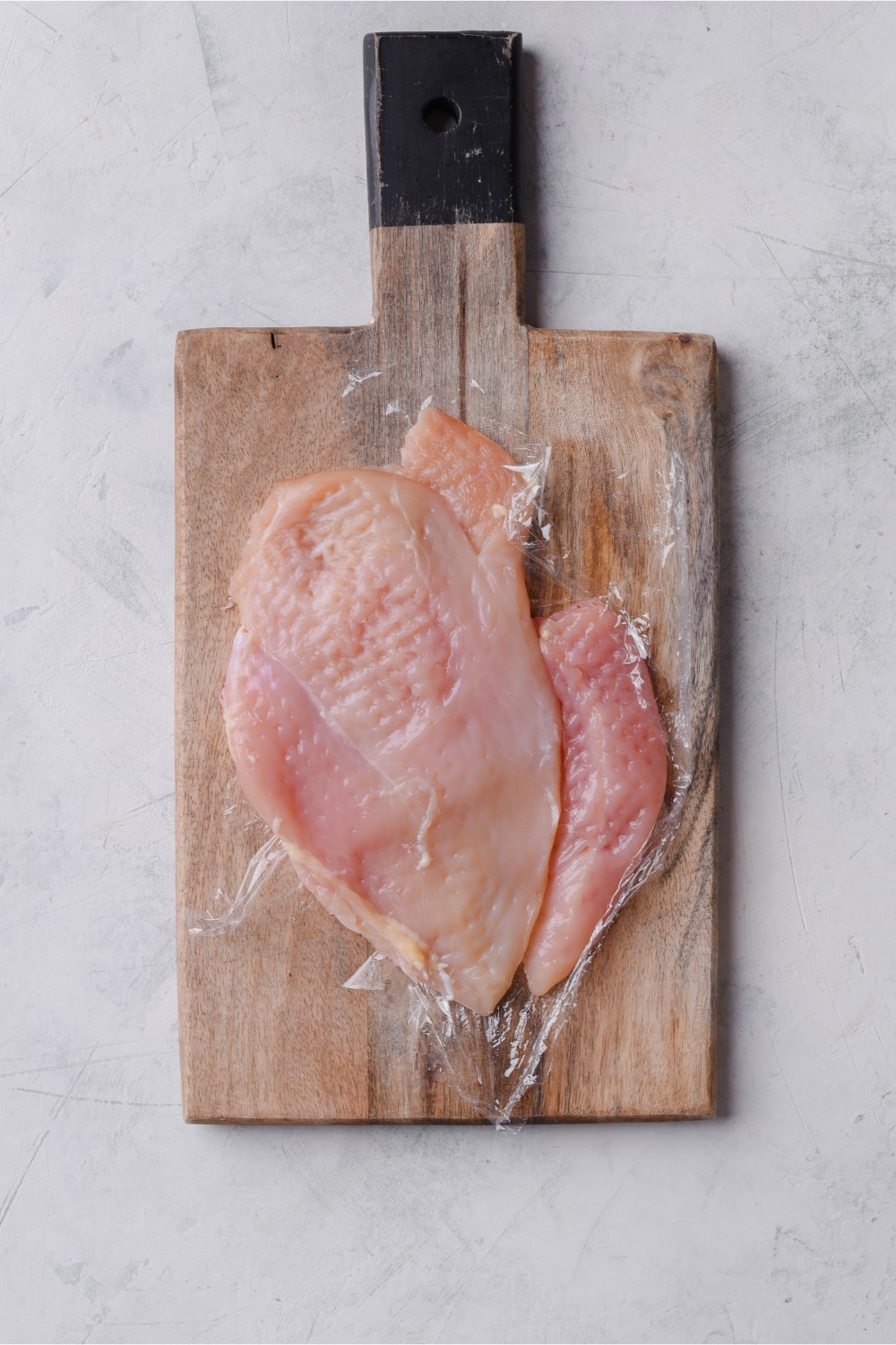 Chicken filets covered with plastic wrap on a wooden cutting board.