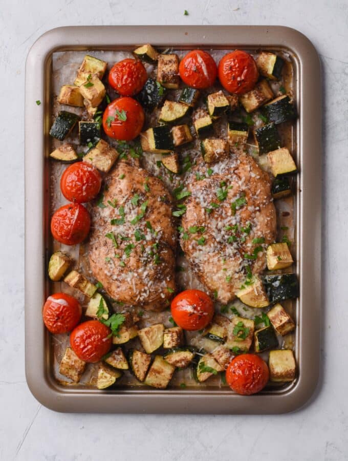 Seasoned baked chicken filets, cherry tomatoes, and zucchini on a sheet pan lined with parchment paper.