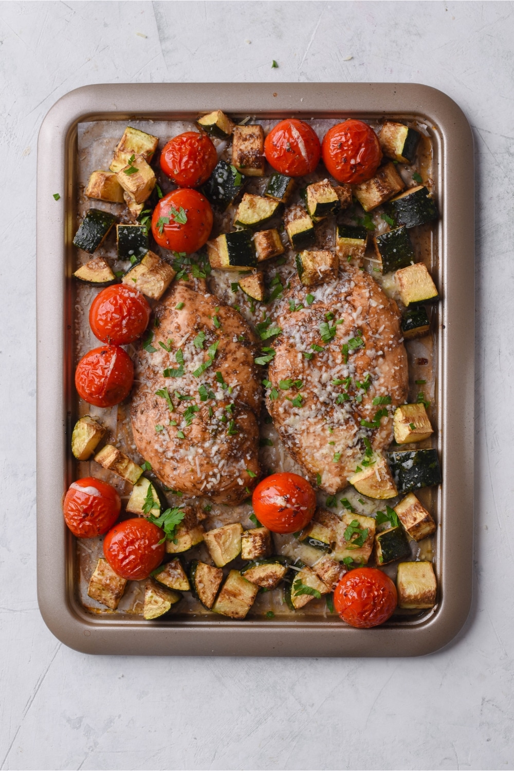 Seasoned baked chicken filets, cherry tomatoes, and zucchini on a sheet pan lined with parchment paper.