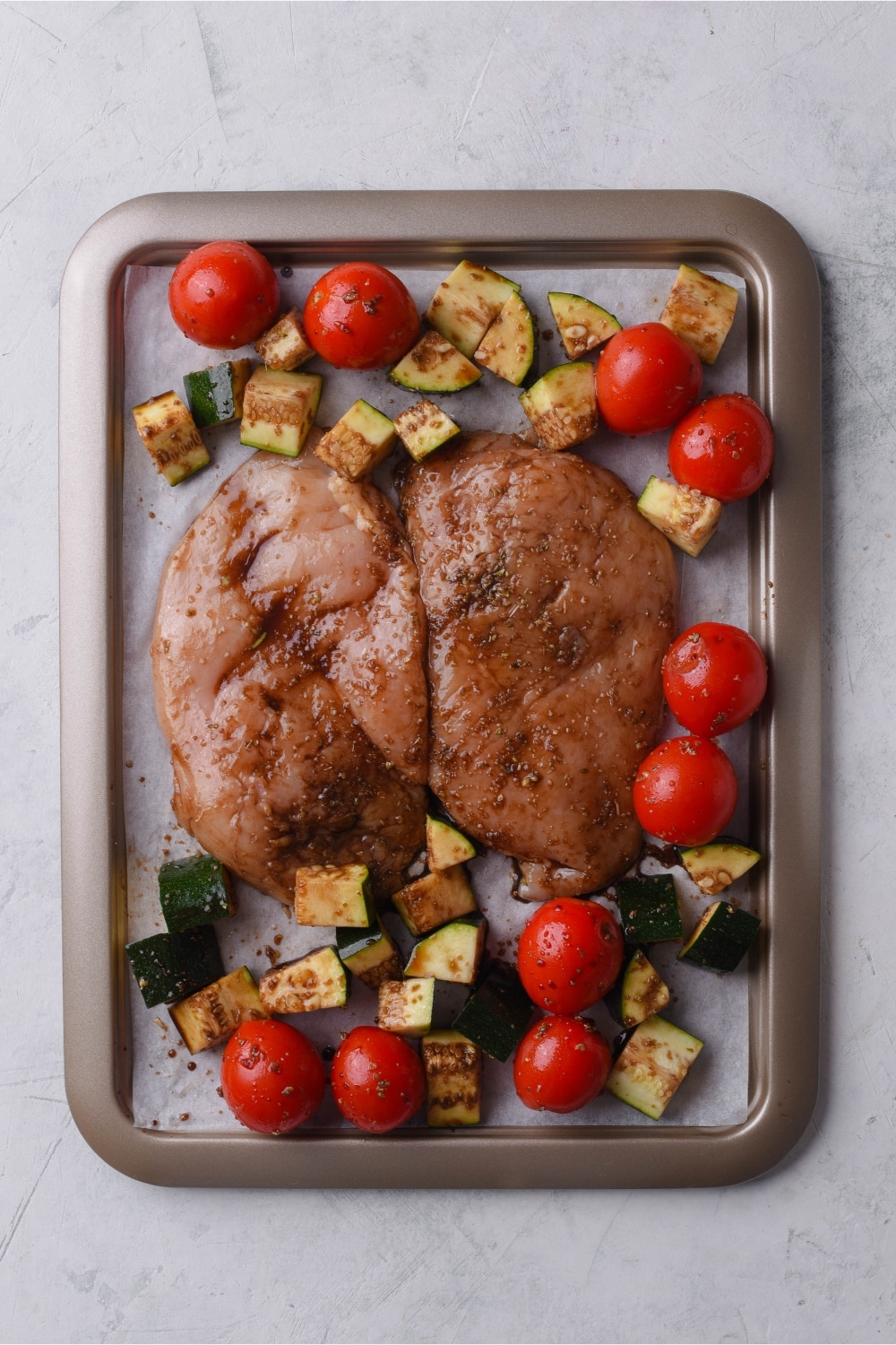 Seasoned uncooked chicken filets, cherry tomatoes, and zucchini on a sheet pan lined with parchment paper.