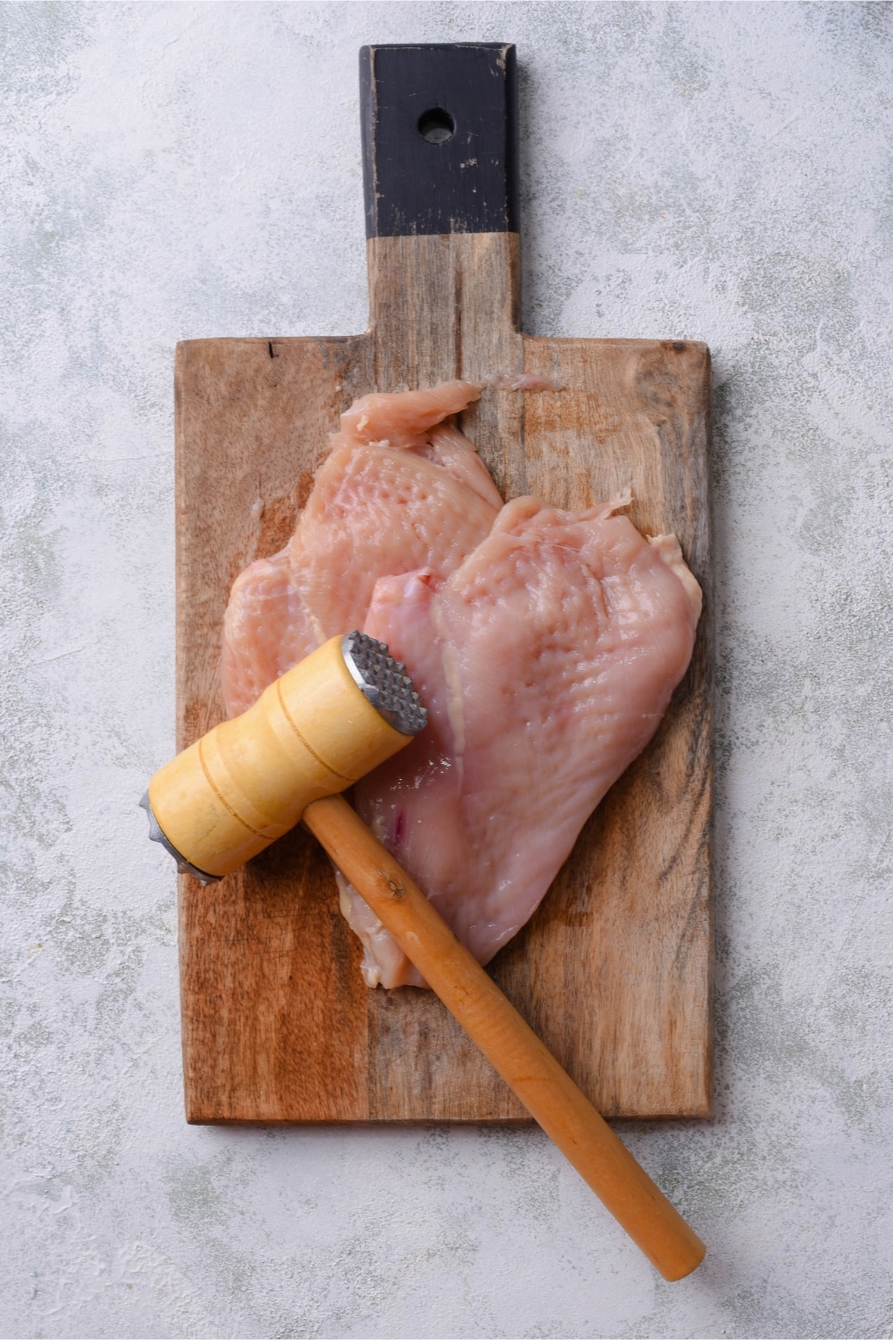 A thin chicken breast on a wooden cutting board with a meat mallet.