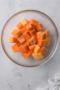 A glass bowl of cubed uncooked butternut squash with seasoning.