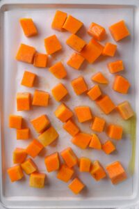 Seasoned uncooked butternut squash spread out on a parchment paper-lined baking sheet.
