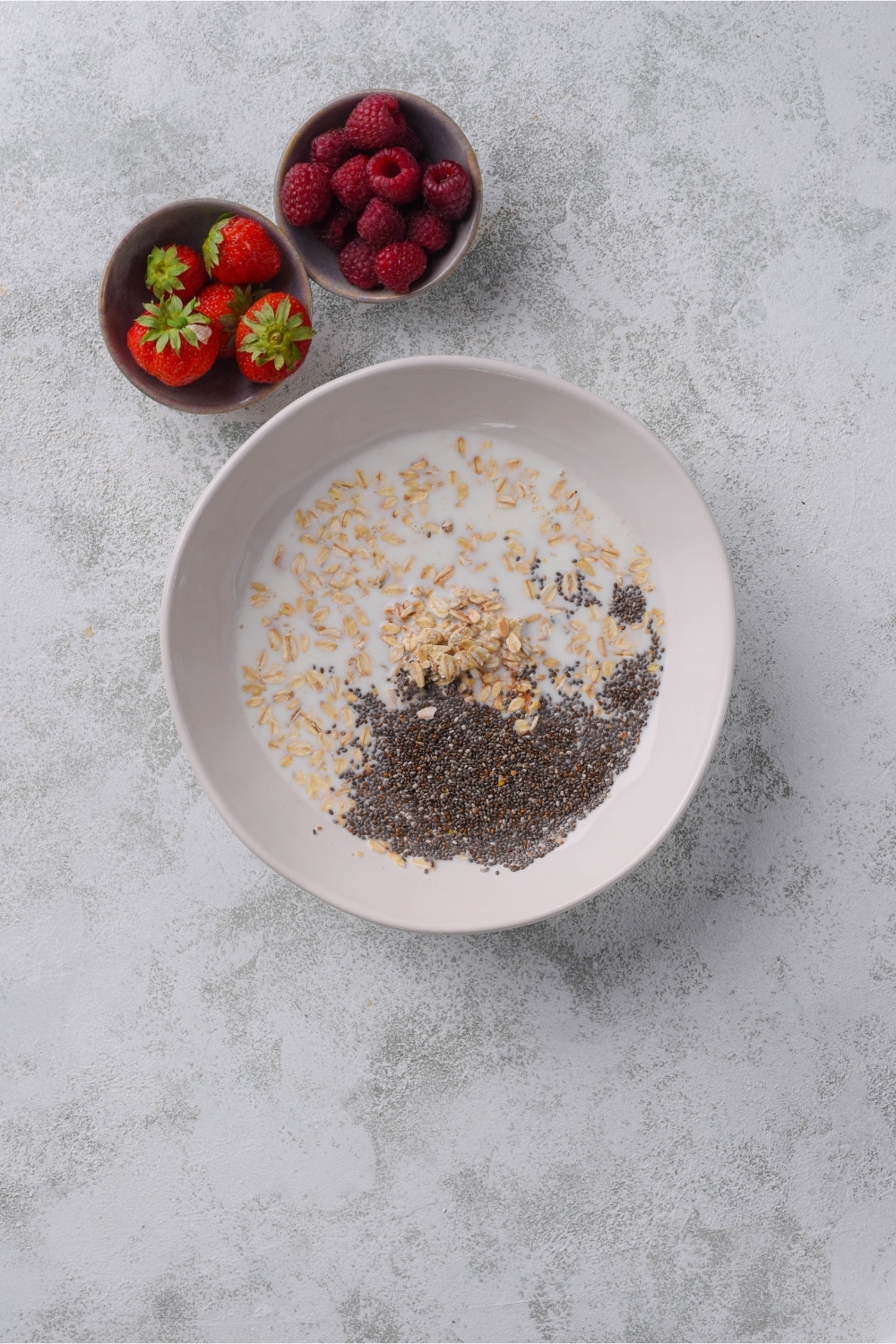 Unmixed overnight oats mixture in a bowl next to bowls of strawberries and raspberries.