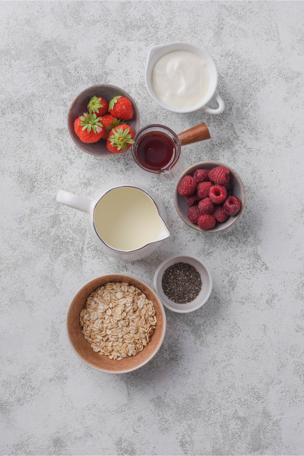 Bowls of greek yogurt, strawberries, raspberries, chia seeds, almond milk, maple syrup, and rolled oats.