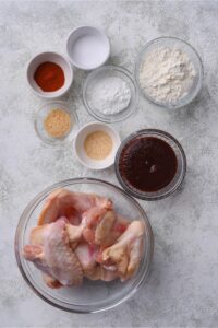 A bowl of paprika, a bowl of flour, a bowl of baking powder, a bowl of bbq sauce, and a bowl of chicken wings.