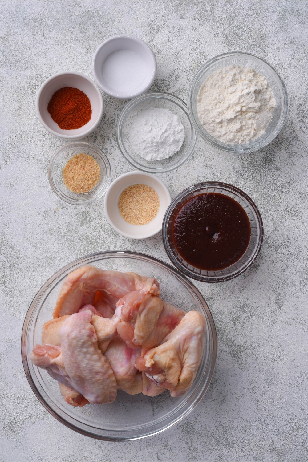 A bowl of paprika, a bowl of flour, a bowl of baking powder, a bowl of bbq sauce, and a bowl of chicken wings.