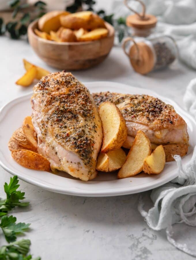 Two seasoned chicken breasts on a plate with potato wedges.