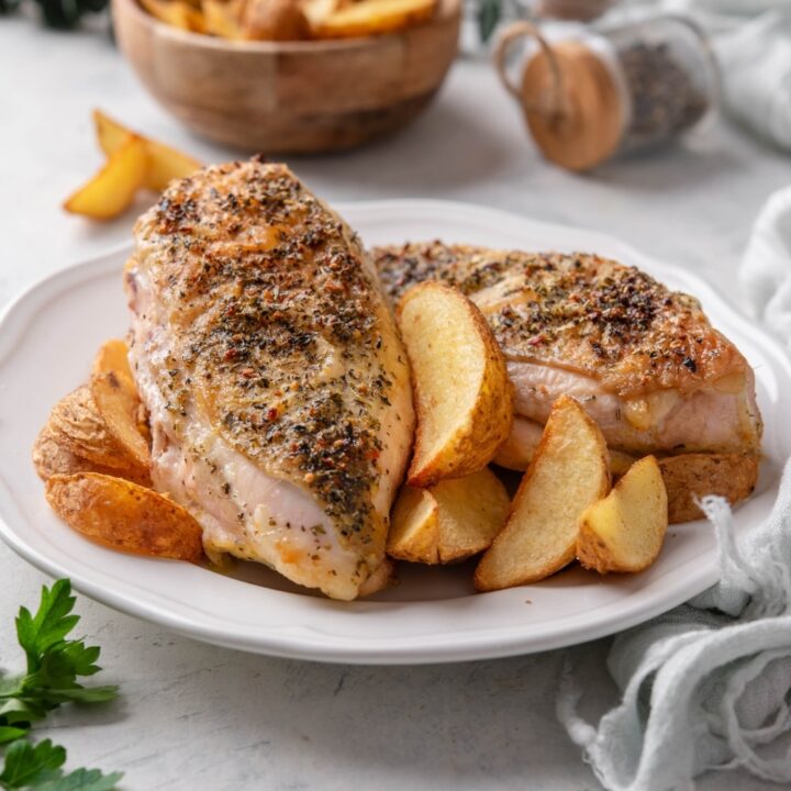 Two seasoned chicken breasts on a plate with potato wedges.