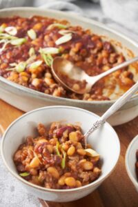 A bowl of beans in sauce with ground beef and green onion. In the background is a baking dish filled with more beans.