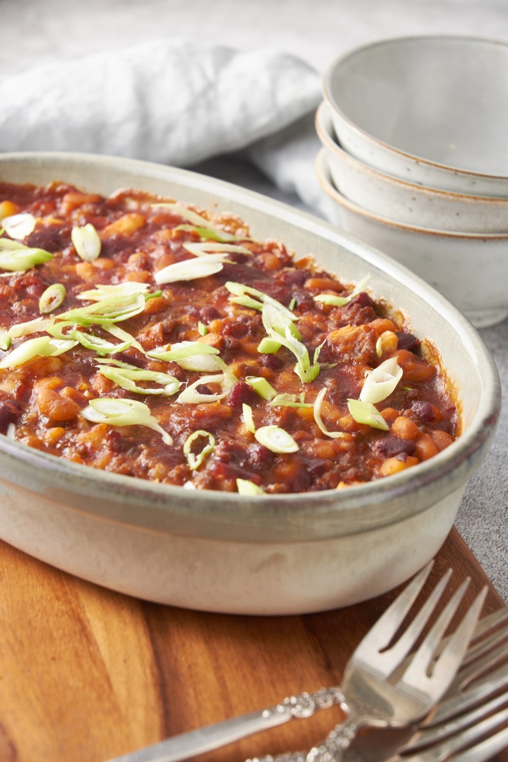 A baking dish filled with baked calico beans garnished with green onion on a wooden board with three forks next to the baking dish.