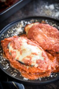 A plate with two chicken breasts covered in red sauce, sliced eggplant, and melted cheese.