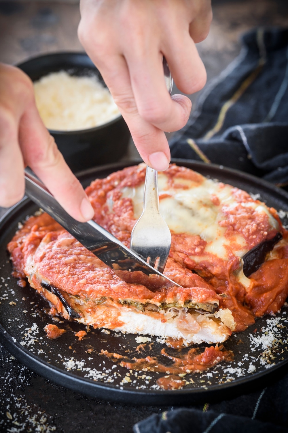 A plate with two chicken breasts covered in red sauce, sliced eggplant, and melted cheese. One chicken breast is being cut using a fork and knife.