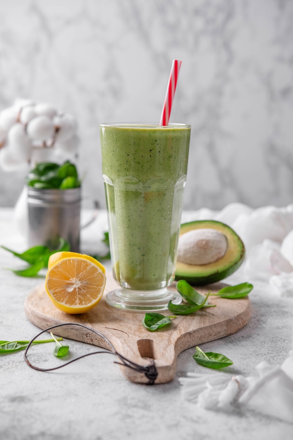A green smoothie in a glass with a straw on a wooden board surrounded by spinach leaves, a halved lemon, and a halved avocado.