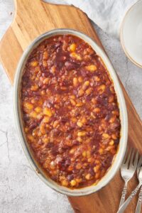 A baking dish filled with baked calico beans on a wooden board with three forks next to the baking dish.