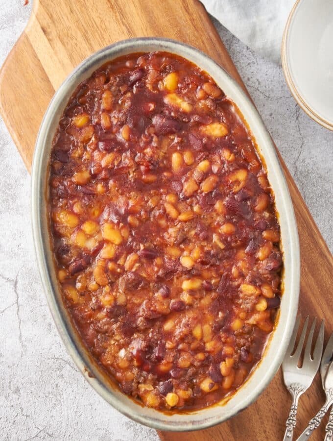 A baking dish filled with baked calico beans on a wooden board with three forks next to the baking dish.