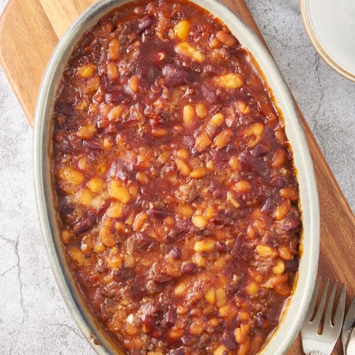 A baking dish filled with baked calico beans on a wooden board with three forks next to the baking dish.