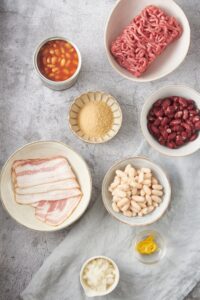 An assortment of ingredients including bowls of raw ground beef, red kidney beans, butter beans, onion, mustard, brown sugar, raw bacon, and an open can of baked beans.