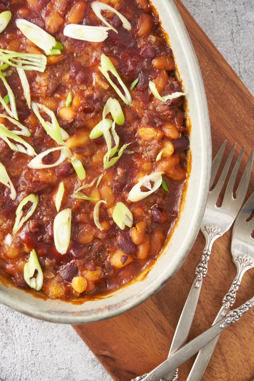 Close up of a baking dish filled with baked calico beans garnished with green onions and three forks are next to the baking dish.