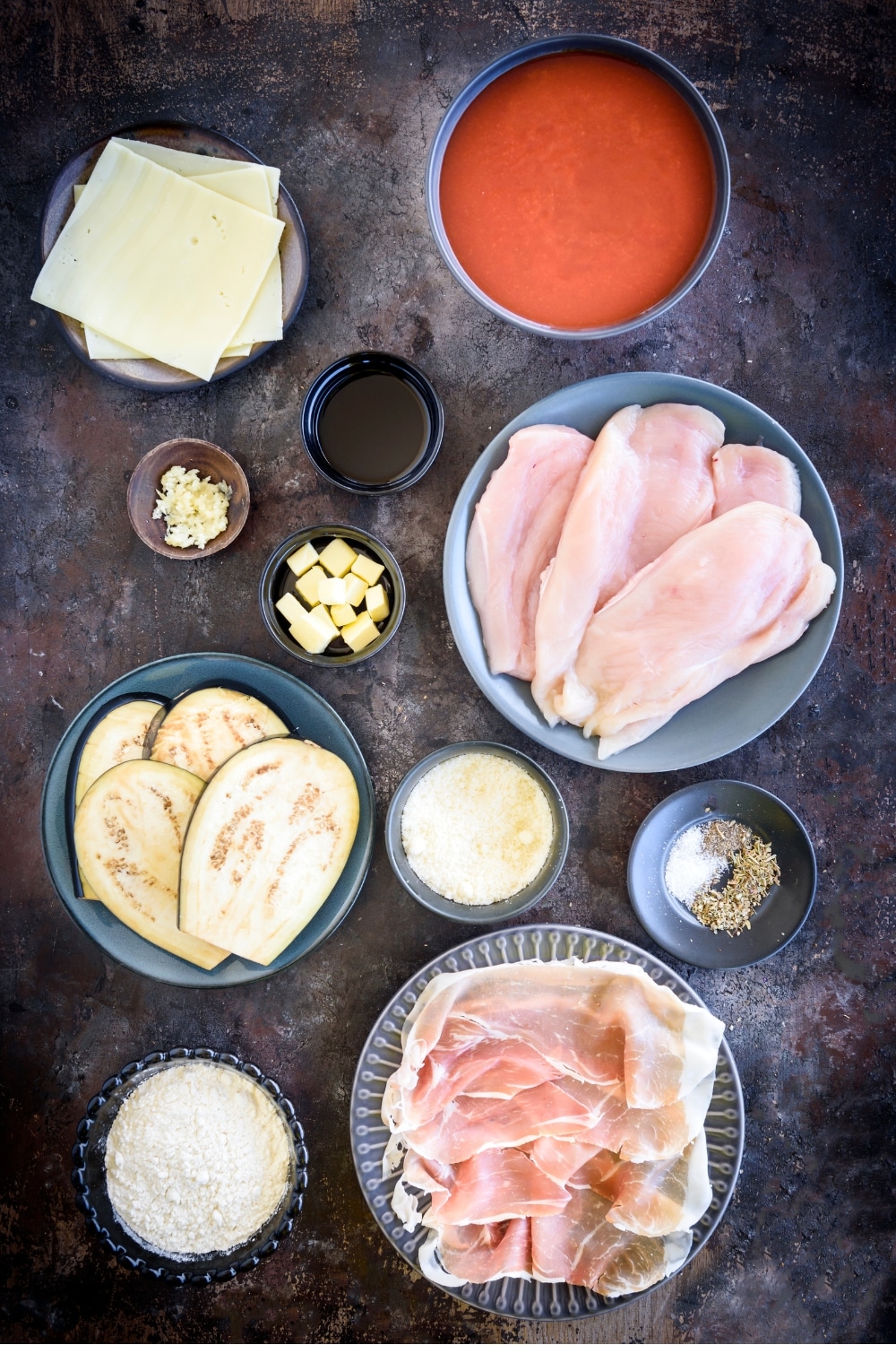 An assortment of ingredients including plates of raw chicken cutlets, sliced prosciutto, sliced eggplant, sliced cheese, and bowls of tomato sauce, flour, butter cubes, oil, and seasonings.