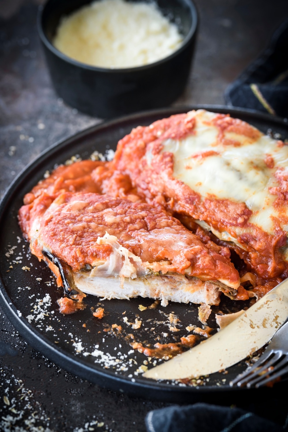 A plate of chicken Sorrentino with one chicken cutlet that has been sliced in half next to another chicken cutlet covered in melted cheese. There is a knife on the plate and in the background is a bowl of parmesan cheese.