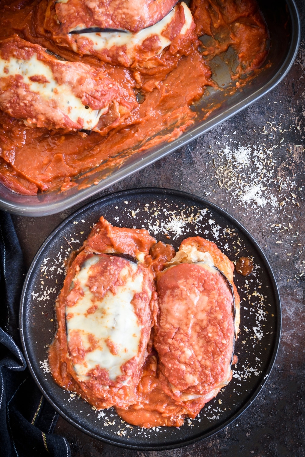 Overhead view of a plate of chicken Sorrentino with two chicken breasts covered in red sauce, sliced eggplant, and melted cheese. Beside the plate of chicken is a baking dish filled with more chicken Sorrentino.