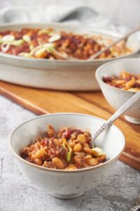 A bowl of calico beans with a spoon in the bowl. In the background is a baking dish filled with more beans and a second serving in another bowl.
