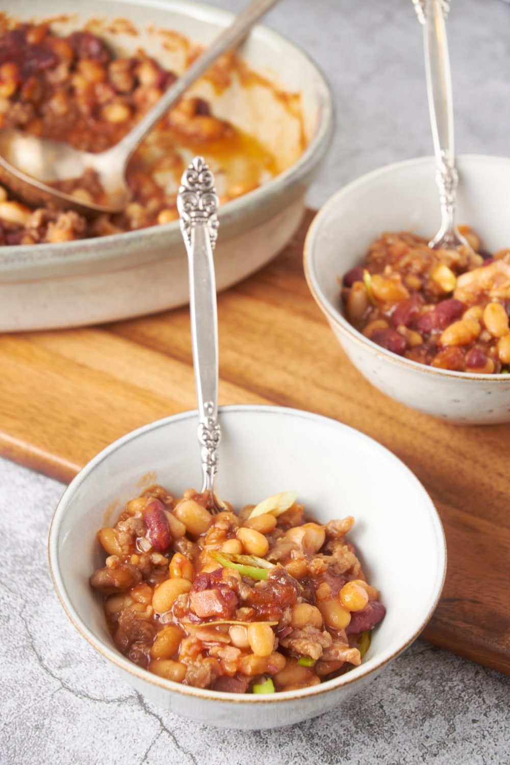 Two bowls of calico beans with spoons in the bowls and a baking dish filled with more beans is in the background.