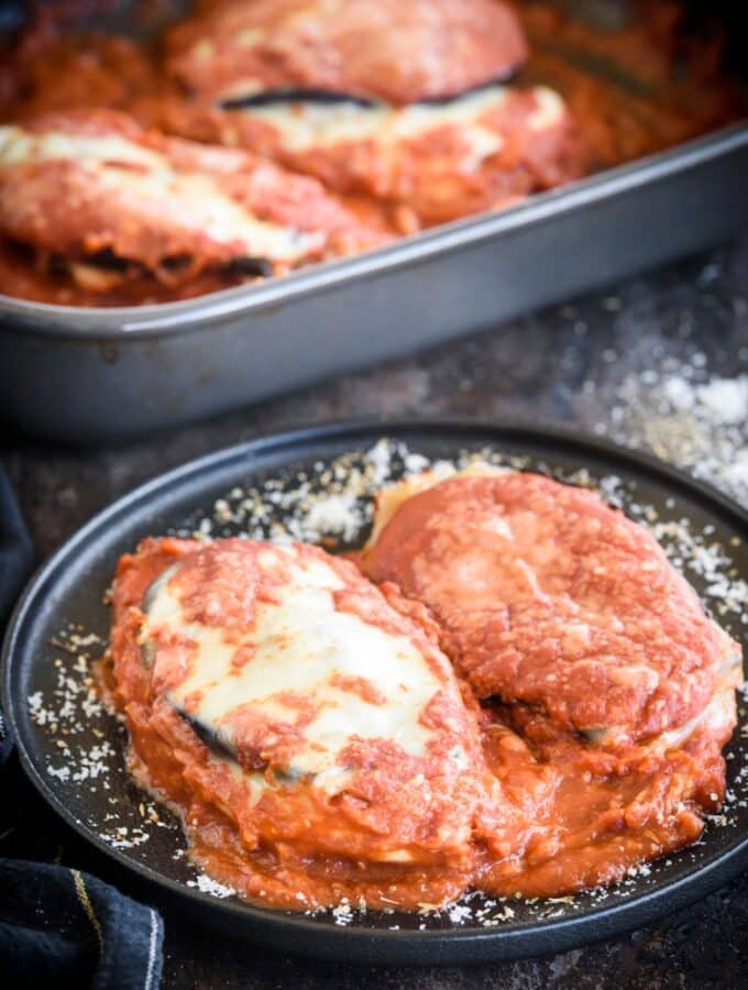 A plate of chicken Sorrentino with two chicken breasts covered in red sauce, sliced eggplant, and melted cheese. Beside the plate of chicken is a baking dish filled with more chicken Sorrentino.