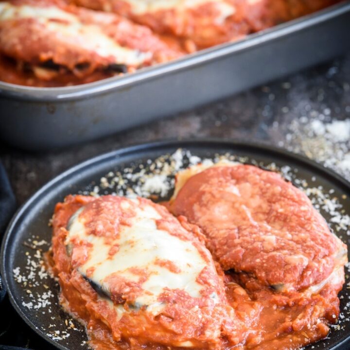 A plate of chicken Sorrentino with two chicken breasts covered in red sauce, sliced eggplant, and melted cheese. Beside the plate of chicken is a baking dish filled with more chicken Sorrentino.