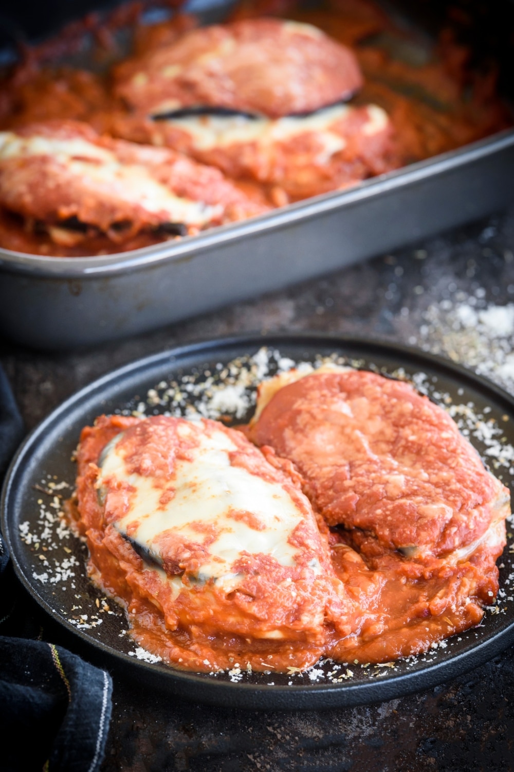 A plate of chicken Sorrentino with two chicken breasts covered in red sauce, sliced eggplant, and melted cheese. Beside the plate of chicken is a baking dish filled with more chicken Sorrentino.