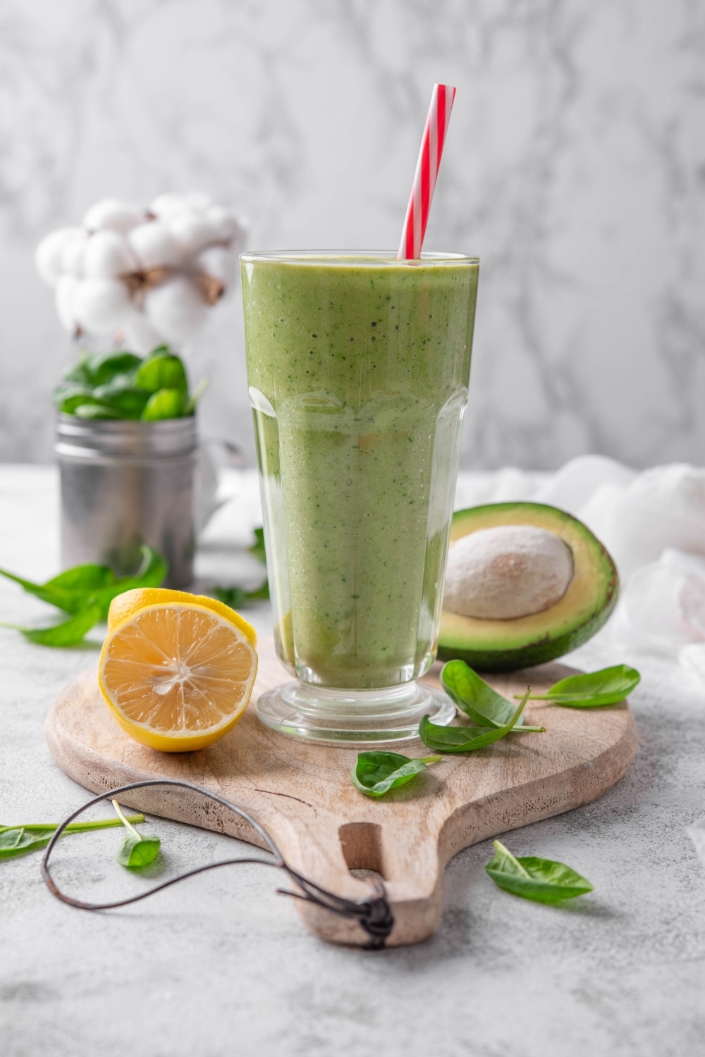 A green smoothie in a glass with a straw on a wooden board surrounded by spinach leaves, a halved lemon, and a halved avocado.