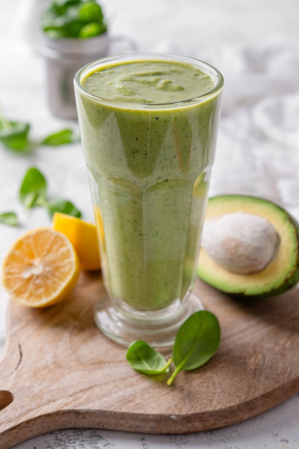 A green smoothie in a tall glass on a wooden board surrounded by spinach leaves, a halved lemon, and a halved avocado.