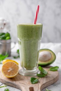 A green smoothie in a glass with a straw on a wooden board surrounded by spinach leaves, a halved lemon, and a halved avocado.