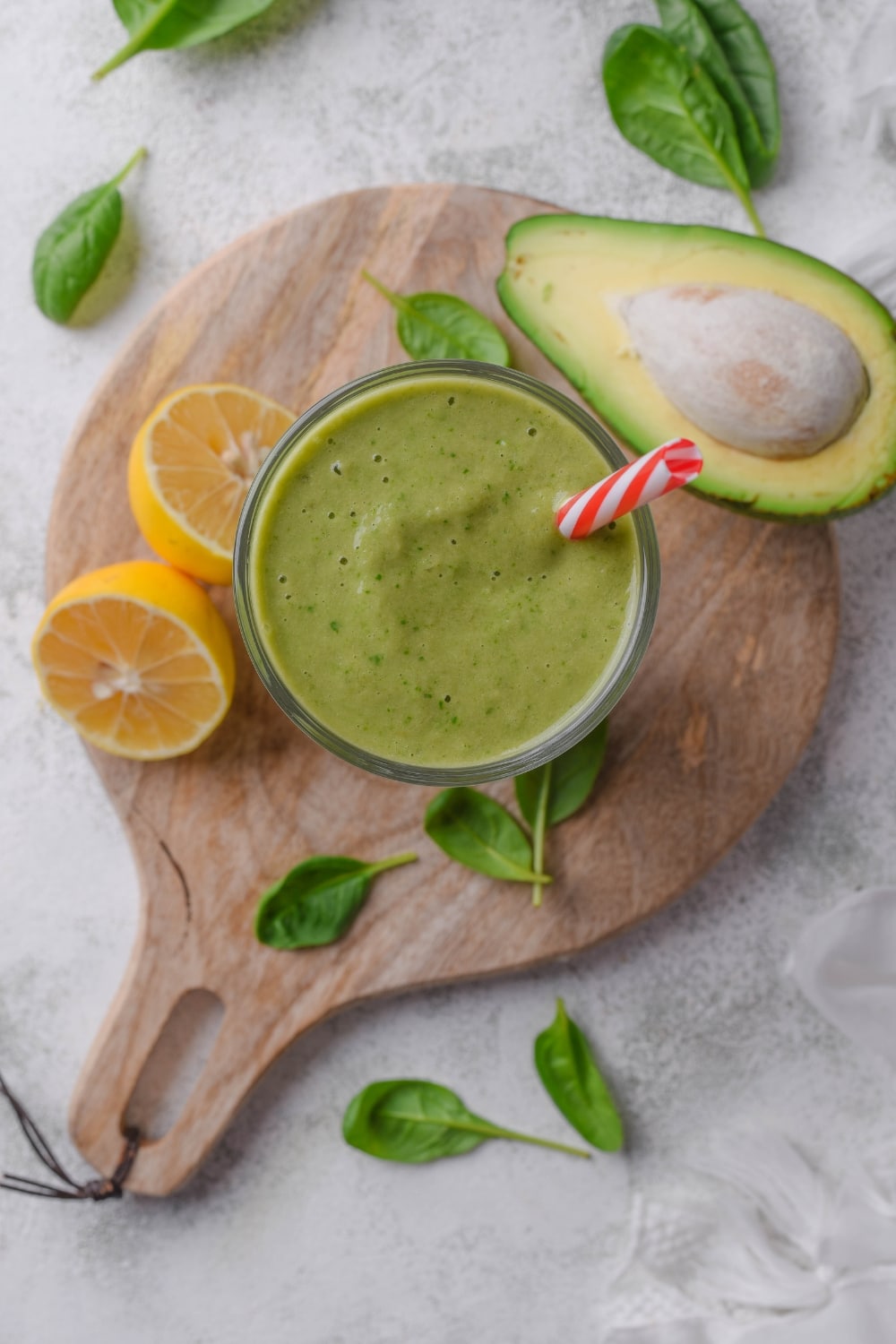 Overhead view of a green smoothie in a glass with a straw in it. The smoothie is surrounded by spinach leaves, a halved lemon, and a halved avocado.