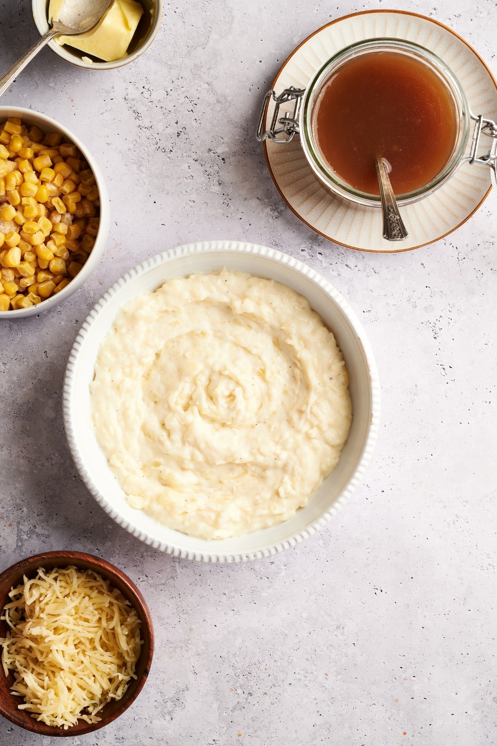 Overhead view of a bowl of creamy mashed potatoes surrounded by bowls of gravy, corn, and shredded cheese.