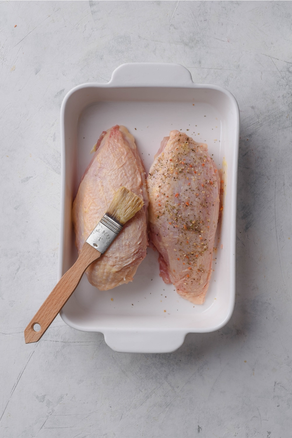 A baking dish with two raw chicken breasts in it. One has been seasoned and one is being brushed with a basting brush.