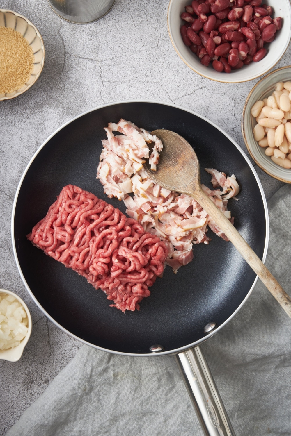 A skillet filled with raw ground beef and bacon. There is a wooden spoon in the skillet.