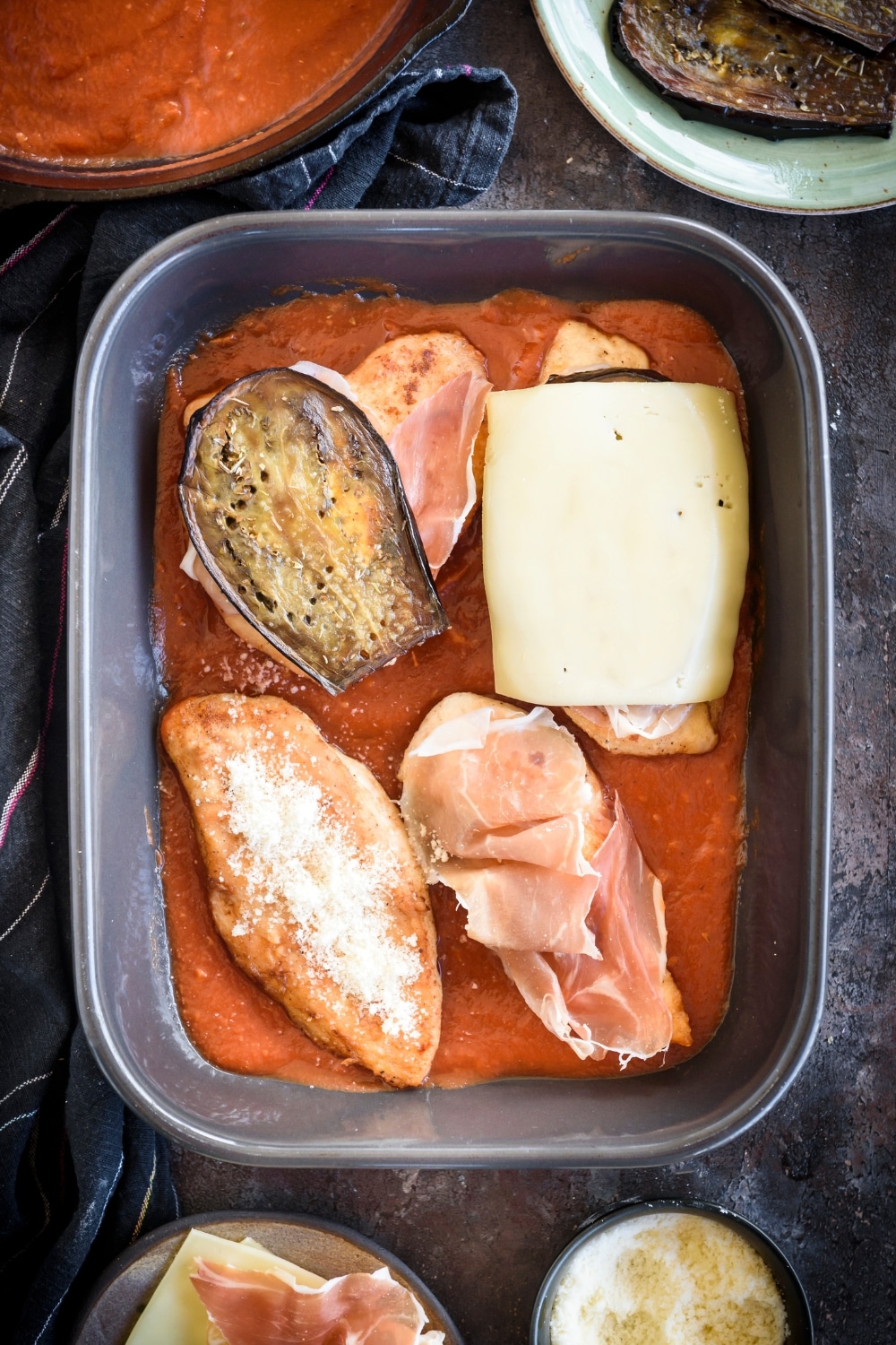 A baking dish filled with tomato sauce and four chicken breasts. One breast is topped with grated cheese, one is topped with prosciutto, one is topped with prosciutto and an eggplant slice, and one is topped with prosciutto, eggplant, and a slice of cheese.