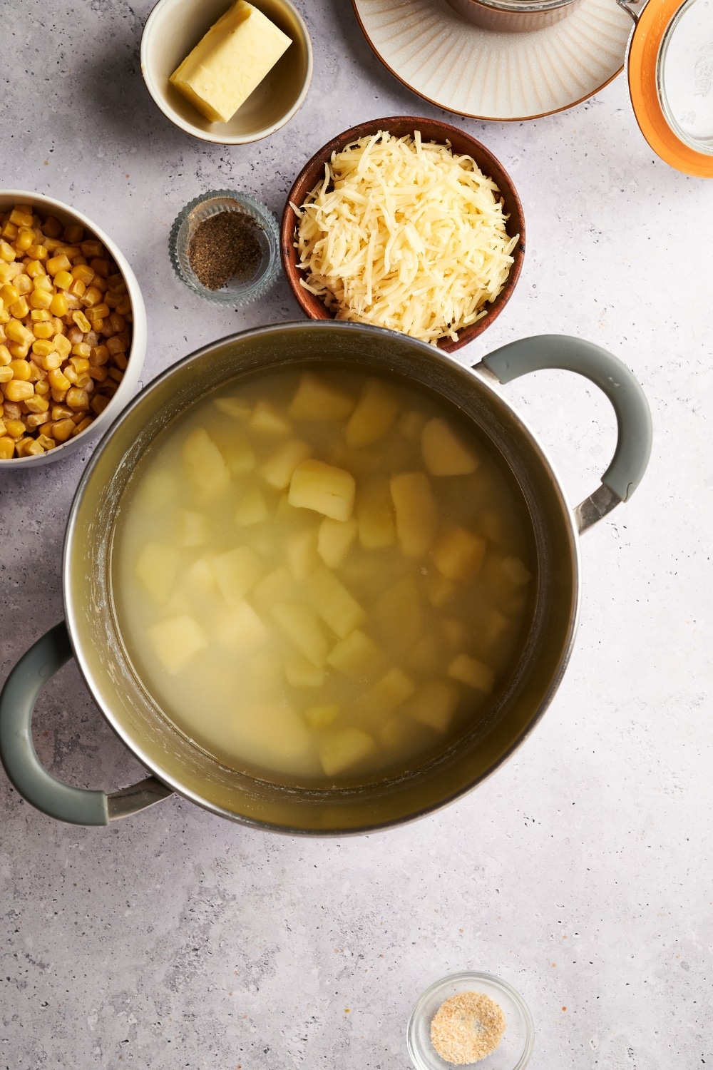 A pot filled with diced potatoes in water. The pot is surrounded by bowls of spices, butter, shredded cheese, and corn.