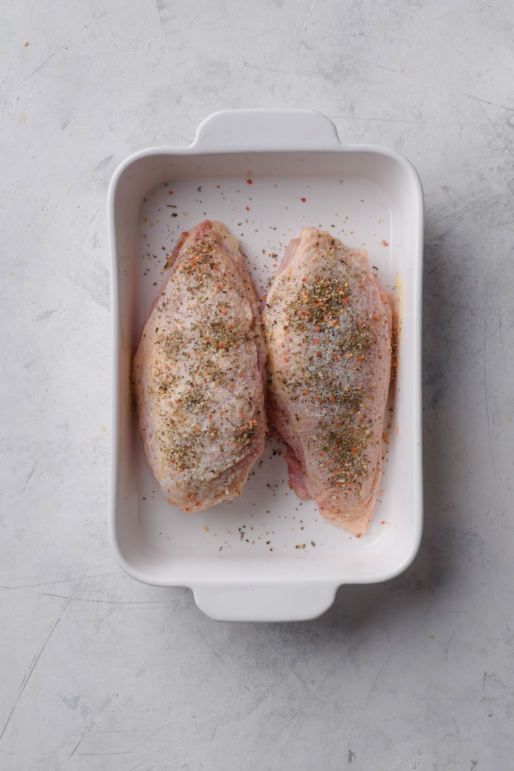 A baking dish with two raw seasoned chicken breasts in it.