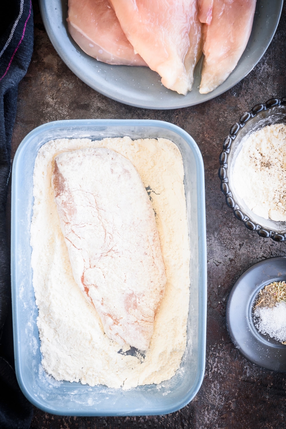 A raw chicken cutlet in a baking dish coated in flour. Next to the baking dish is a bowl of flour, a bowl of spices, and a plate of raw chicken cutlets.