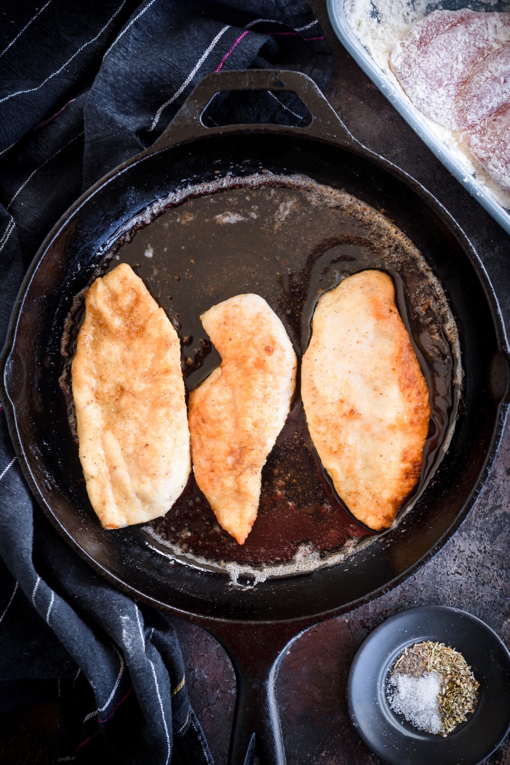 A cast iron skillet with three chicken cutlets frying in oil.