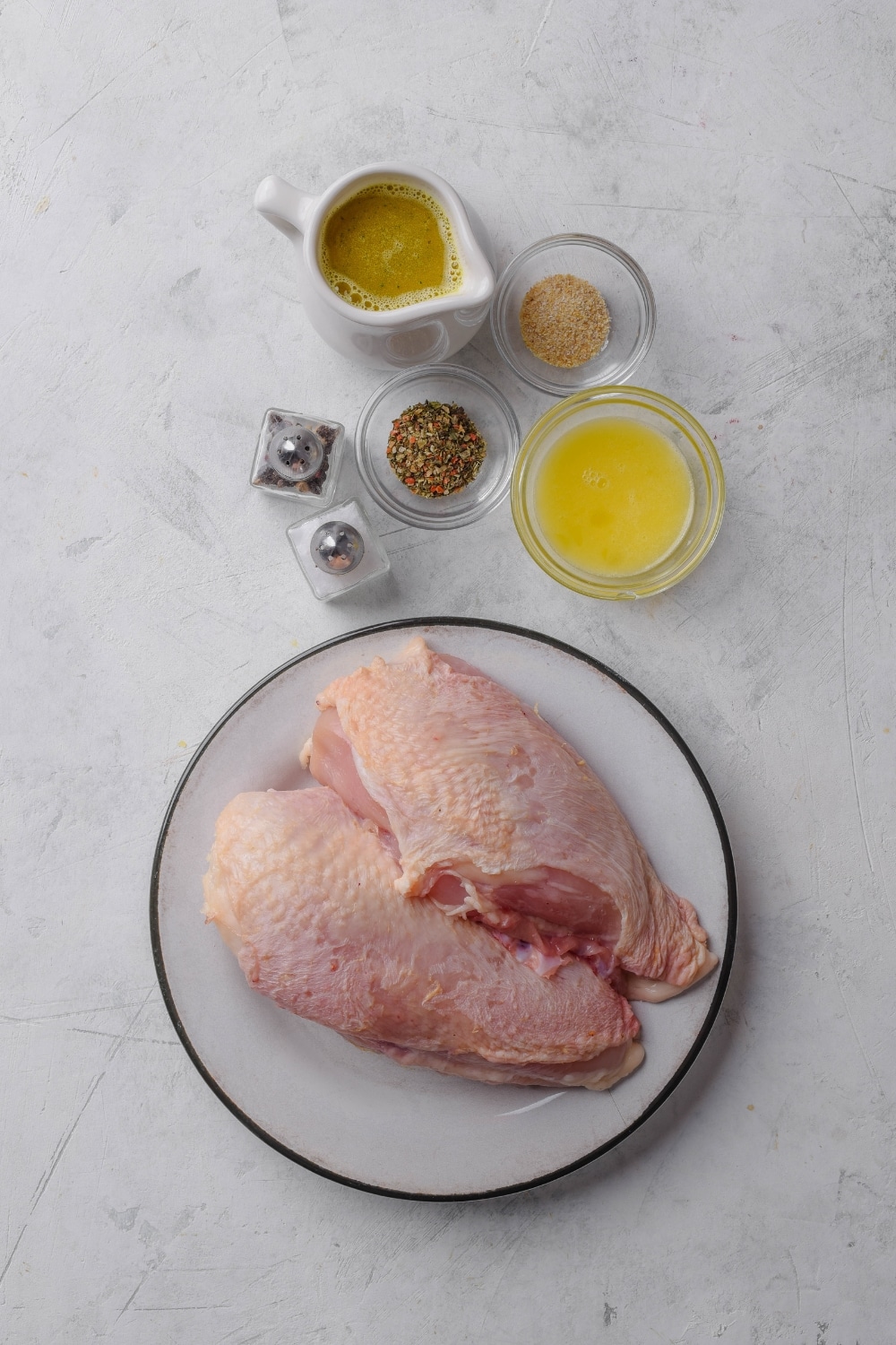 A plate with two raw chicken breasts next to a pitcher of broth, a bowl of butter, salt and pepper shakers, and two bowls of spices.