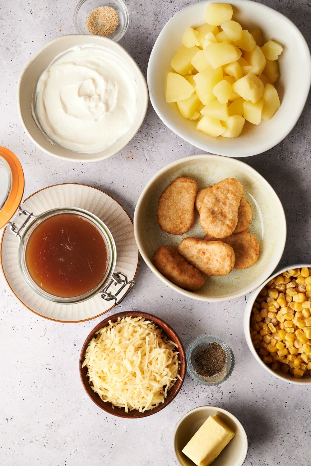 An assortment of ingredients including bowls of diced potatoes, chicken nuggets, brown gravy, cream cheese, shredded cheese, corn, butter, and spices.