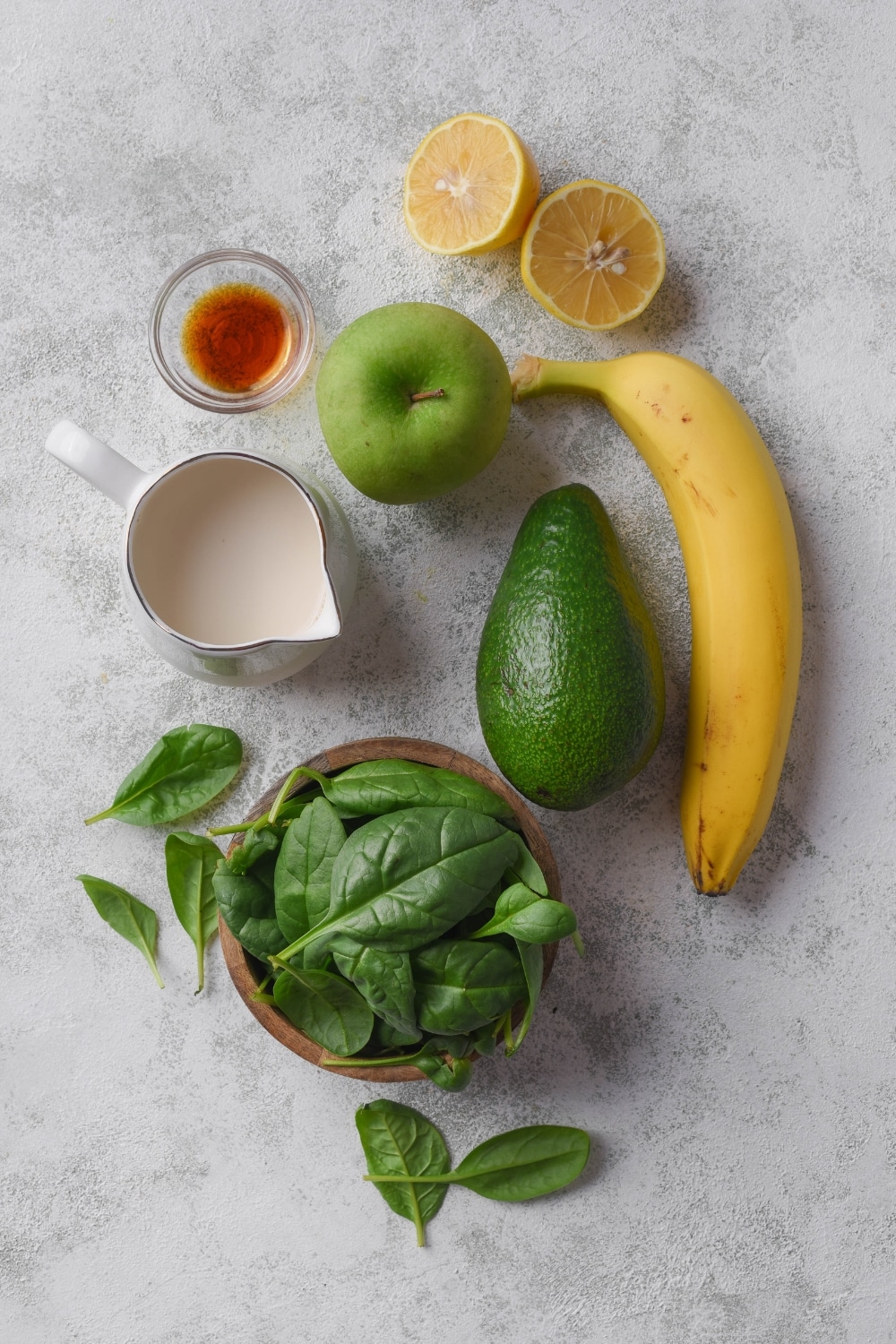 An assortment of ingredients including a bowl of fresh spinach, a pitcher of milk, a bowl of vanilla extract, and a green apple, a halved lemon, a banana, and an avocado.